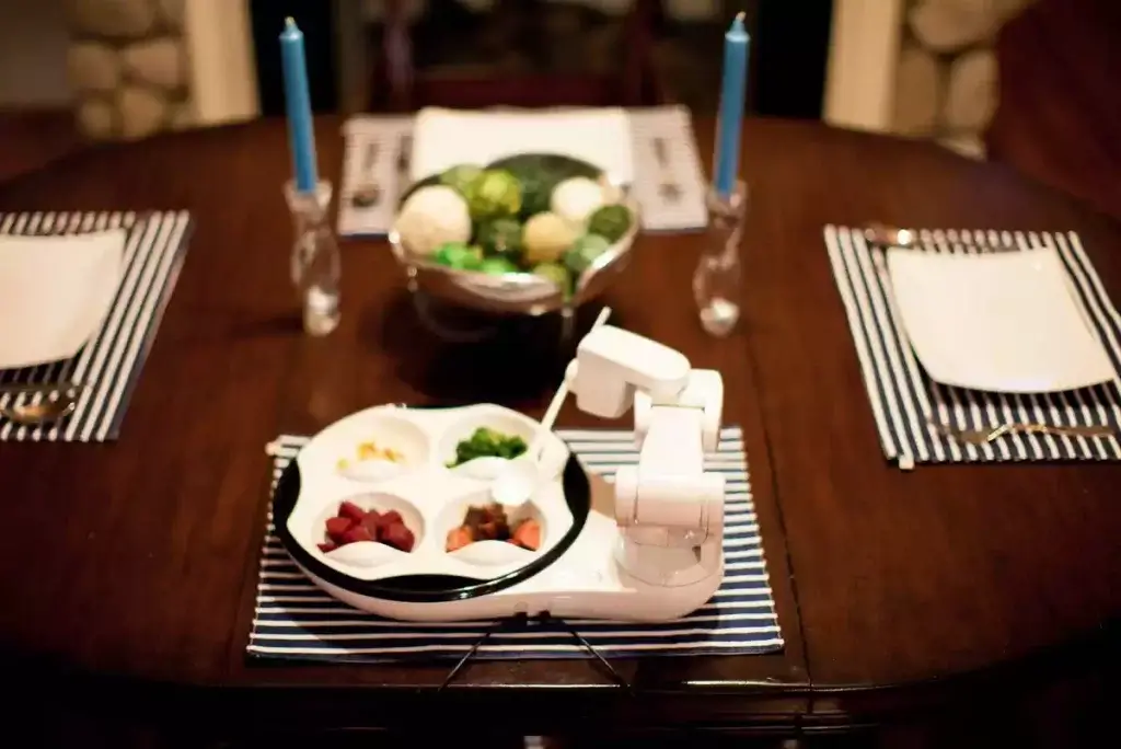De foto toont het robotische eetapparaat Obi op een eettafel thuis, genomen door de schrijver.